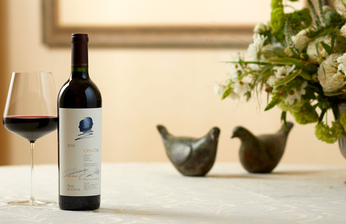 A bottle of Opus One 2019 with two dove statues and green and white florals in the background