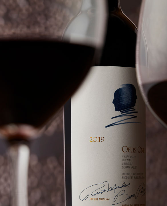 A bottle of Opus One 2019 with two glasses of red wine, and an artistic background
